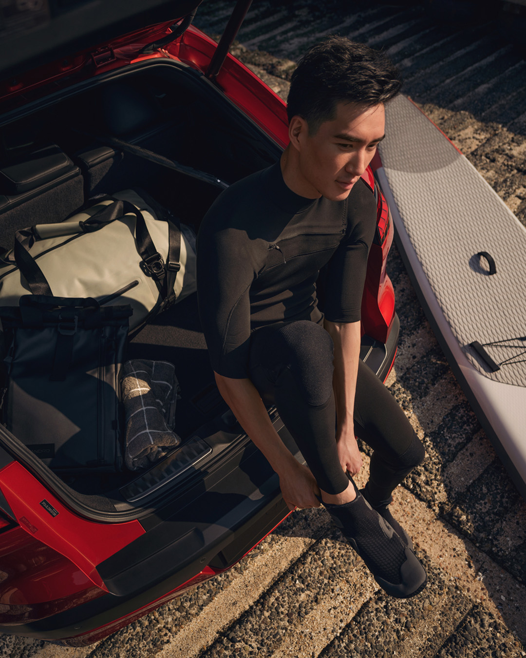 INFINITI driver preparing for surf session beside red crossover SUV, with board and bag visible