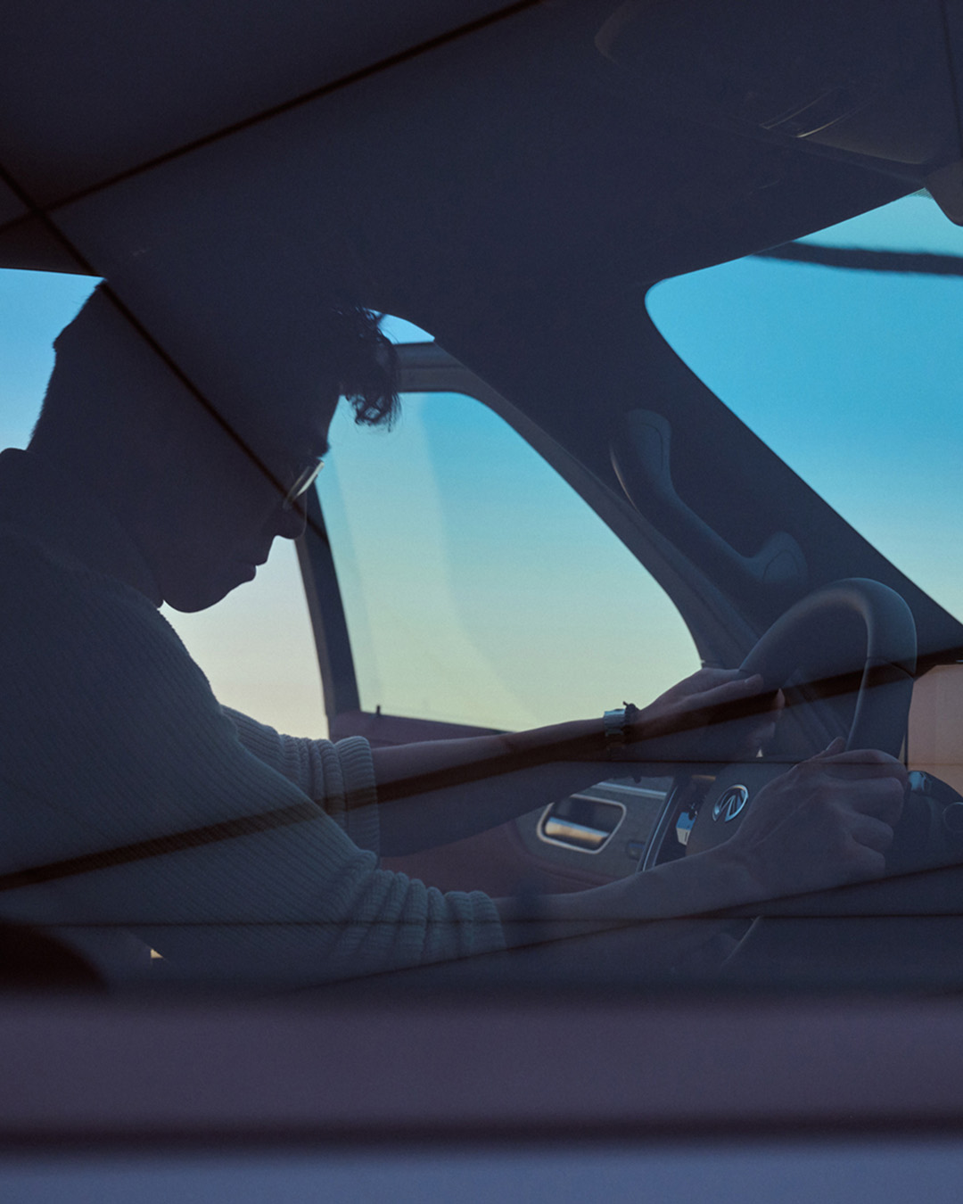 INFINITI driver in profile through passenger glass at dusk, silhouetted against blue sky